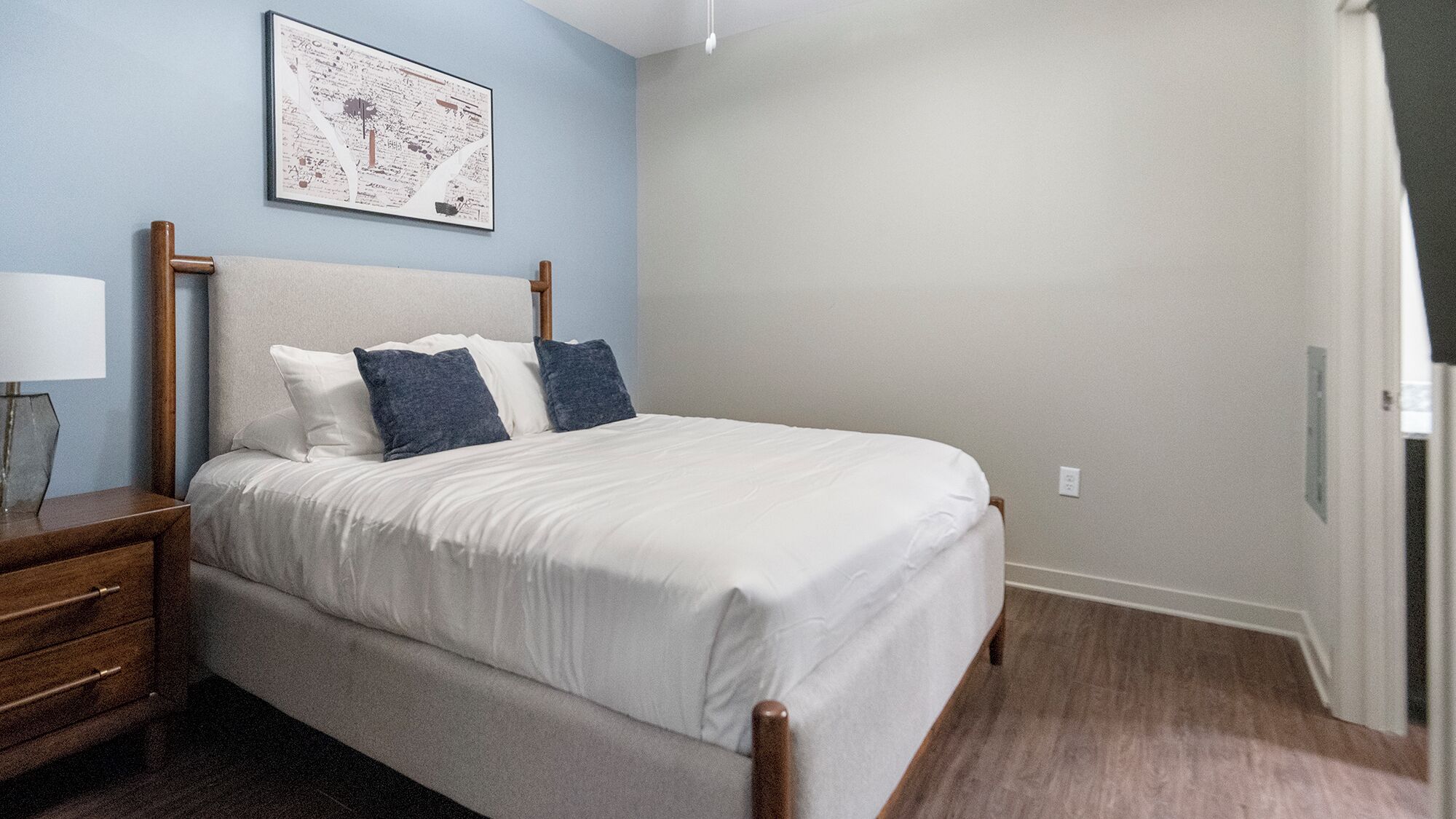 A neatly made bed with white linens and two pillows, a wooden nightstand with a lamp, and a framed map on the wall.