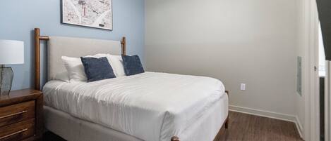 A neatly made bed with white linens and two pillows, a wooden nightstand with a lamp, and a framed map on the wall.