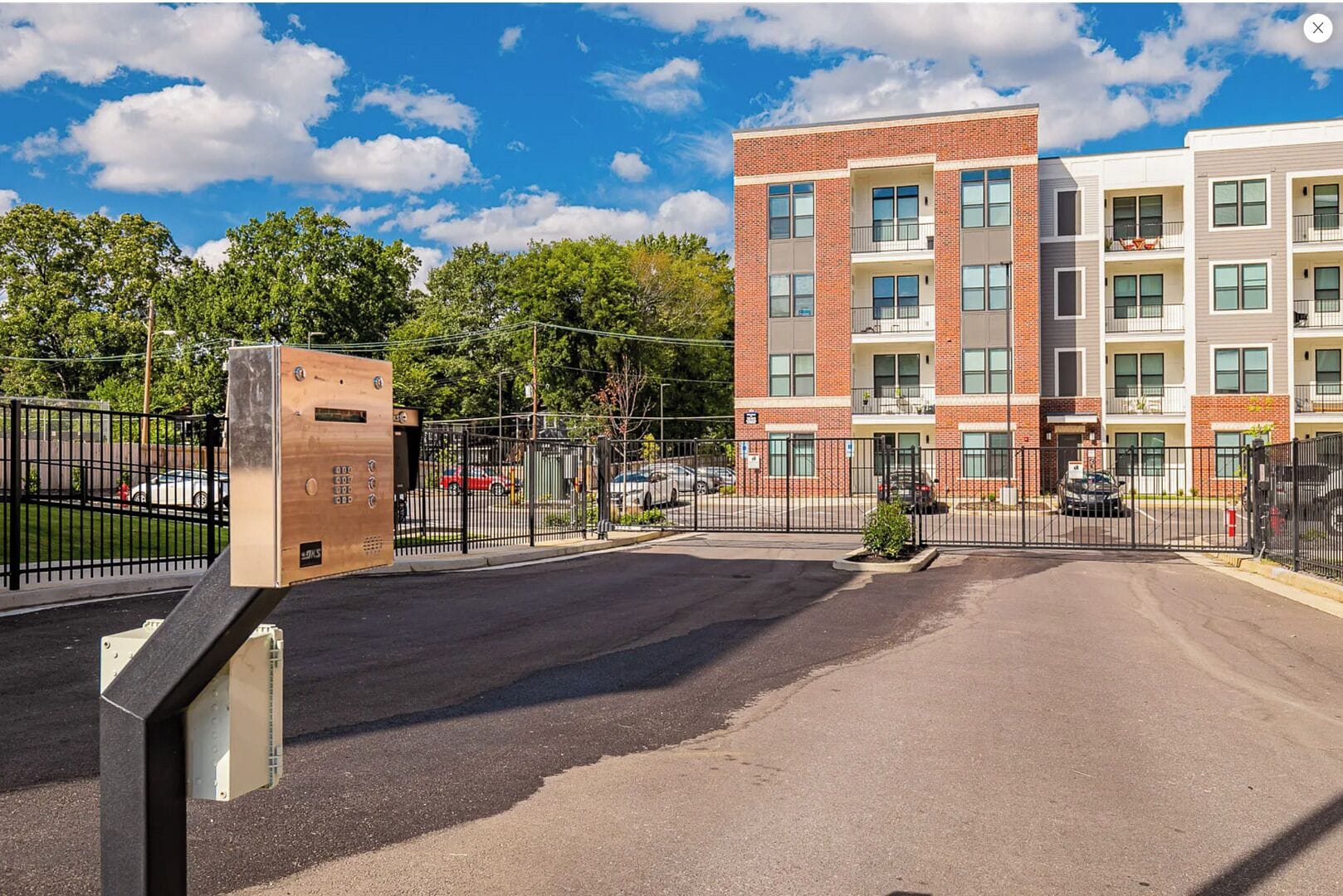A multi-story apartment building with a brick facade, multiple balconies, and a gated parking area.