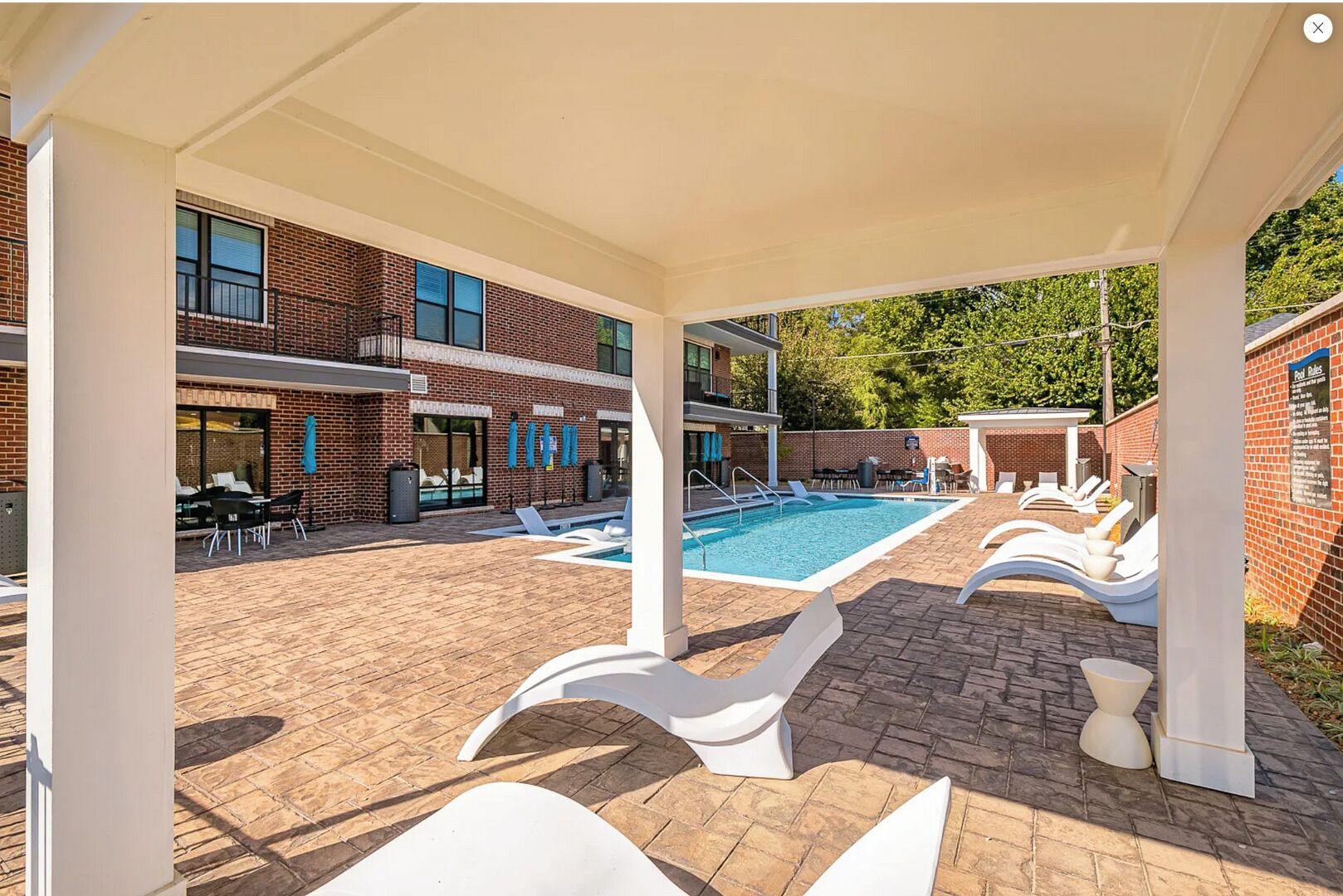 A covered outdoor swimming pool area with lounge chairs and a brick building in the background.