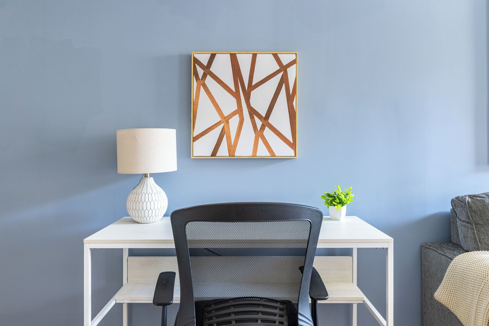 Ein modernes Zimmer mit Schreibtisch, Stuhl, Lampe, Pflanze und Wandbild.