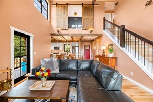 A spacious living area with a black leather sofa, a wooden coffee table, and a vase of flowers. A dining area is visible in the background with a red door.