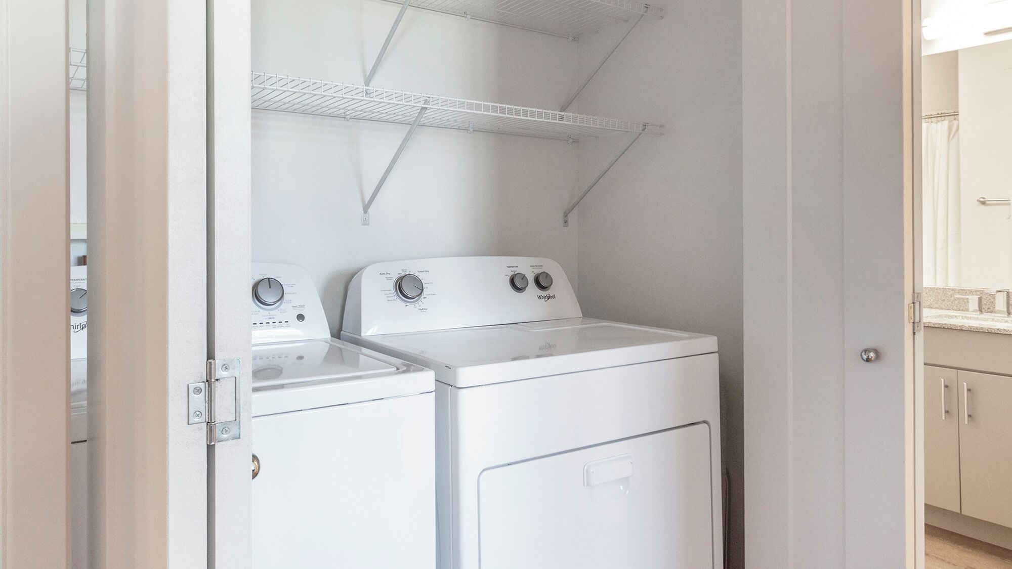A compact laundry room with a front-loading washer and dryer, metal shelving above, and a door leading to a bathroom.