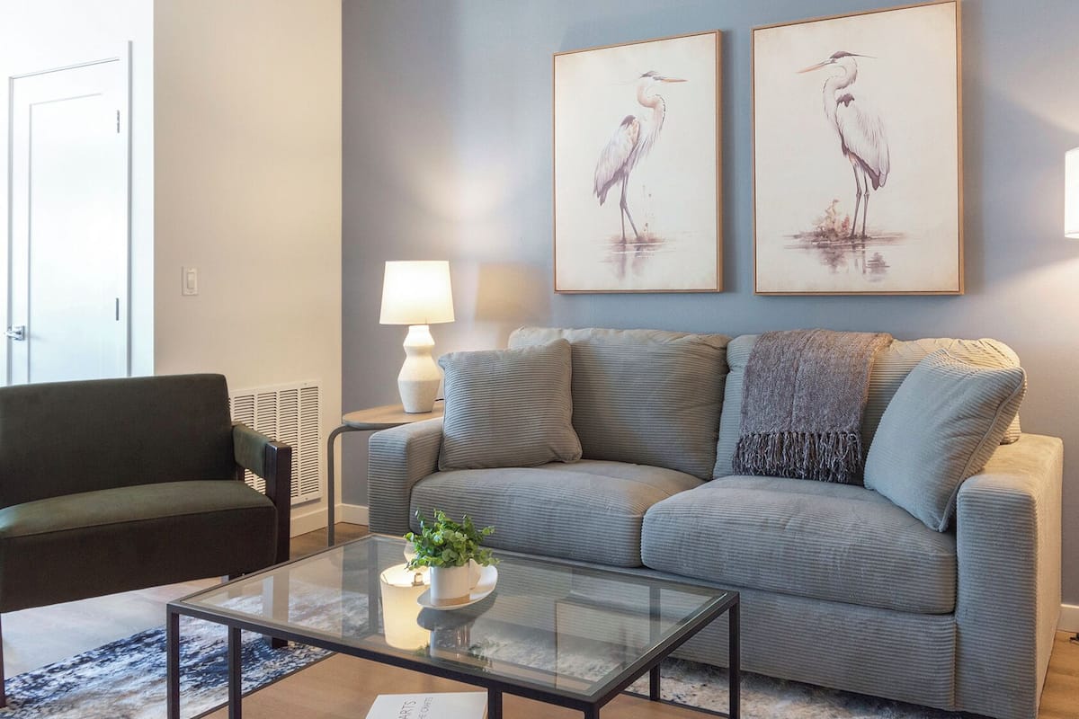 A living room with a grey sofa, a glass coffee table, a lamp, and two framed wall art pieces.
