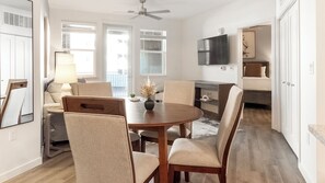 A modern kitchen with a dining area, wooden cabinets, a flat-screen TV, and a large window with blinds.