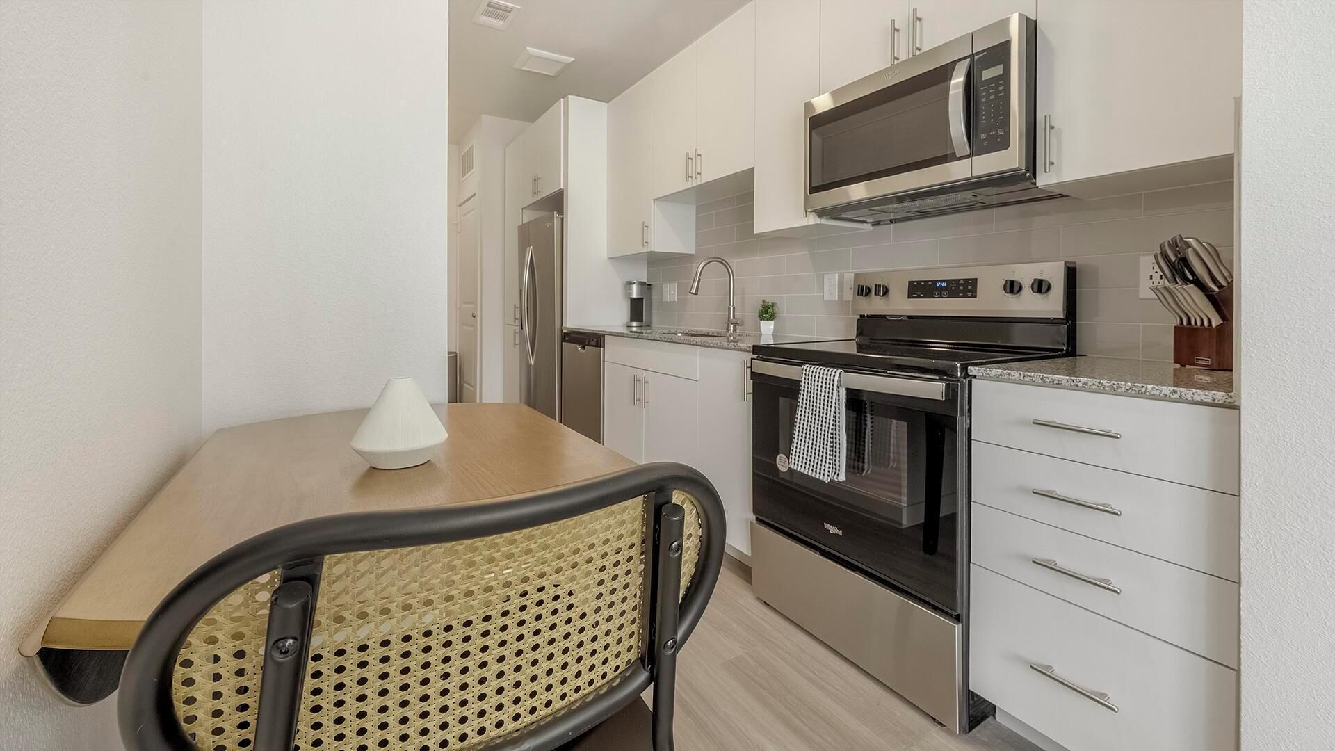 A modern kitchen with white cabinets, stainless steel appliances, a wooden table, and a woven chair.