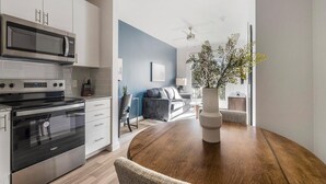 Cozinha moderna com eletrodomésticos de aço inoxidável, sala de jantar com mesa de madeira e sala de estar com sofá e planta.