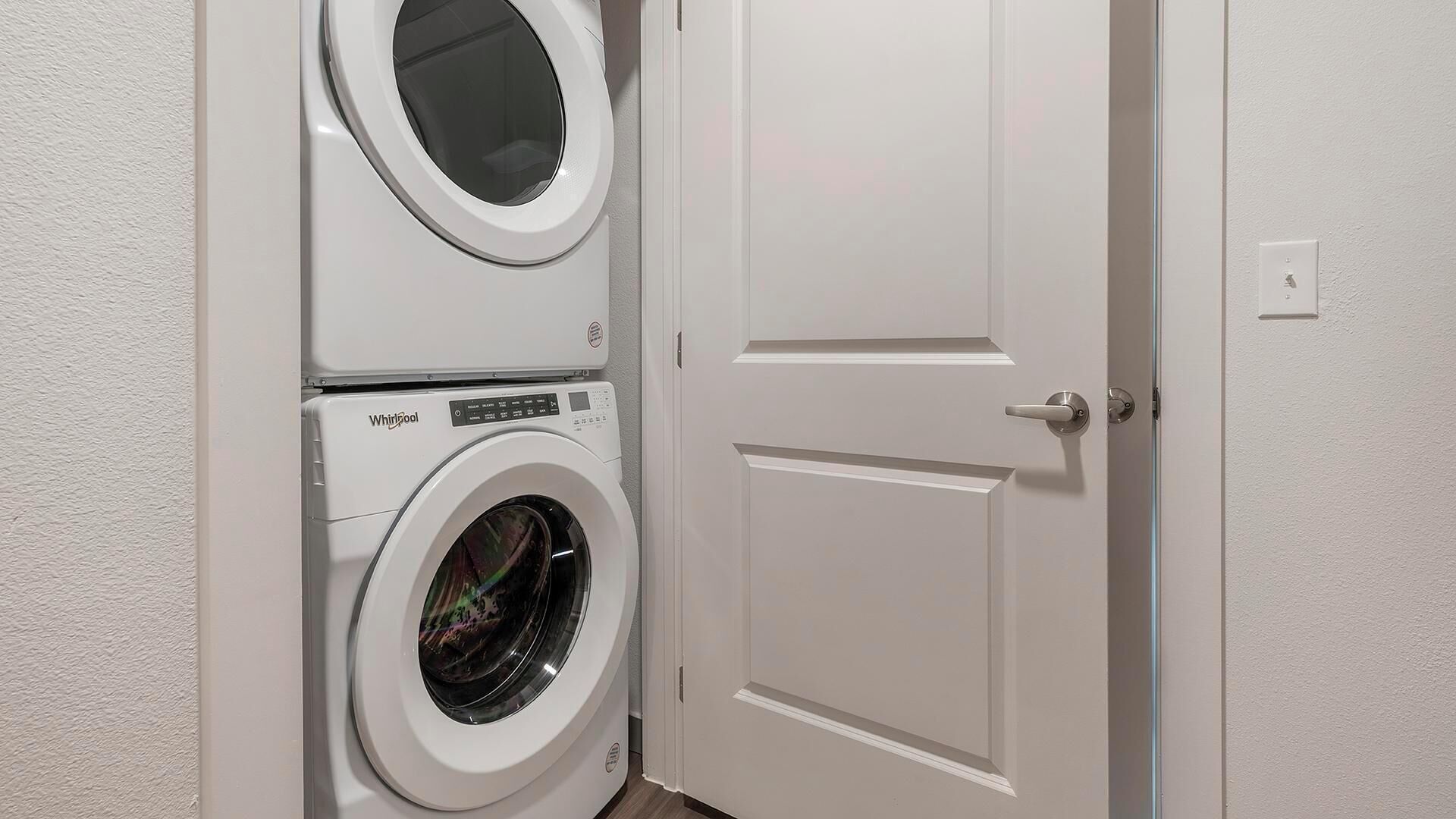 A laundry room with a stacked washer and dryer, a white door, and a light switch on the wall.