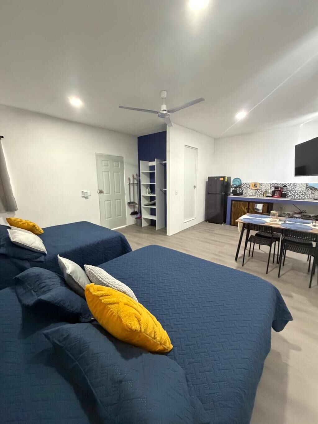A bedroom with two beds, a ceiling fan, a small kitchenette, and a dining area.