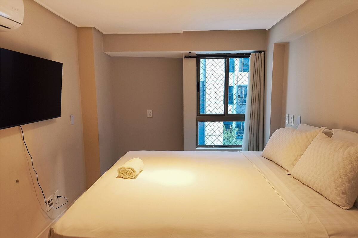 Quarto de hotel com uma cama, televisão na parede, janela com cortinas e uma tomada na parede.