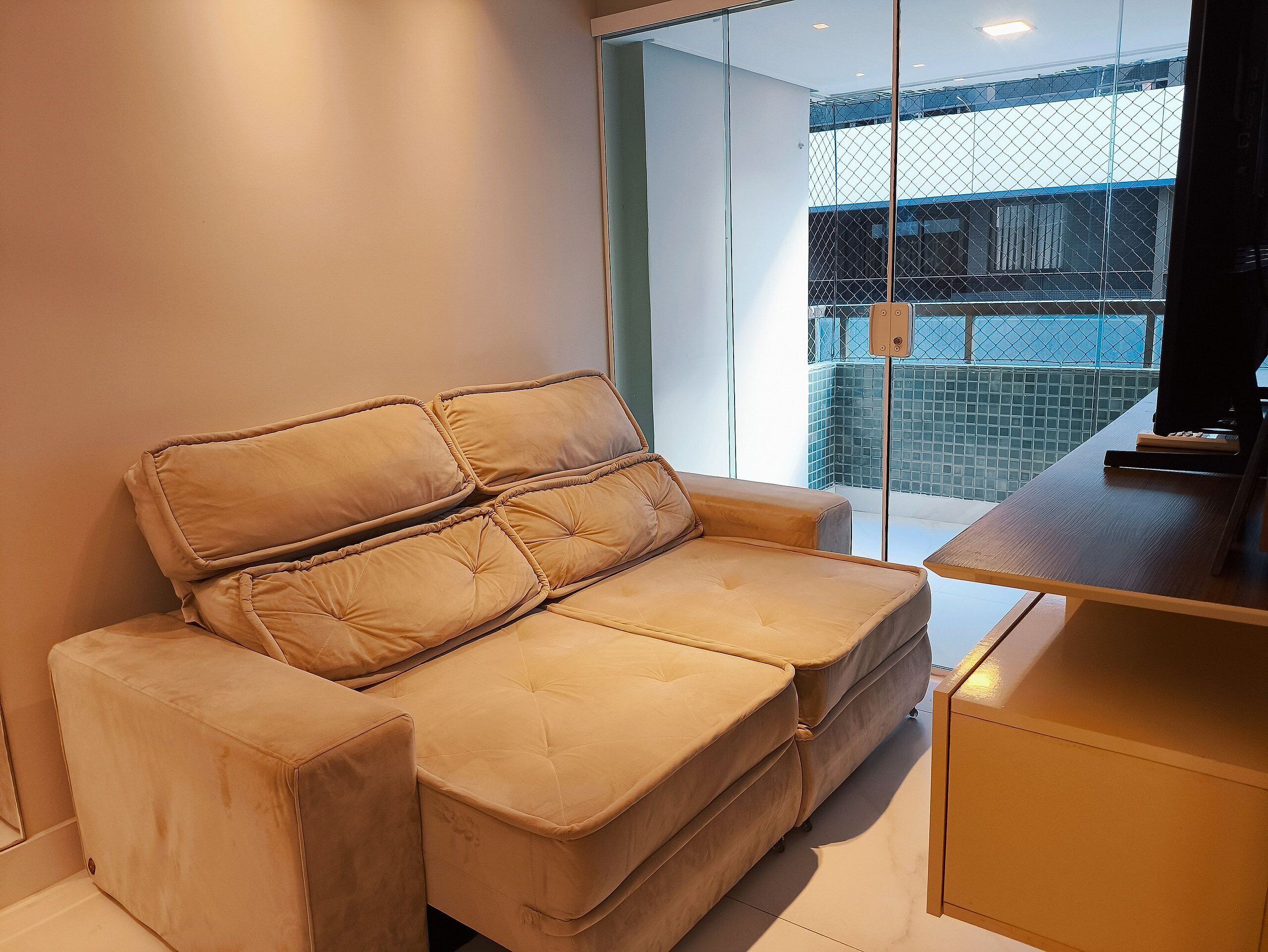A beige sofa with a cushioned backrest, a wooden side table, and a view of a building through a glass door.