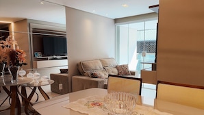 A modern living room with a sofa, a dining table with a glass centerpiece, and a TV mounted on the wall. - Luxor Tambaú (João Pessoa)