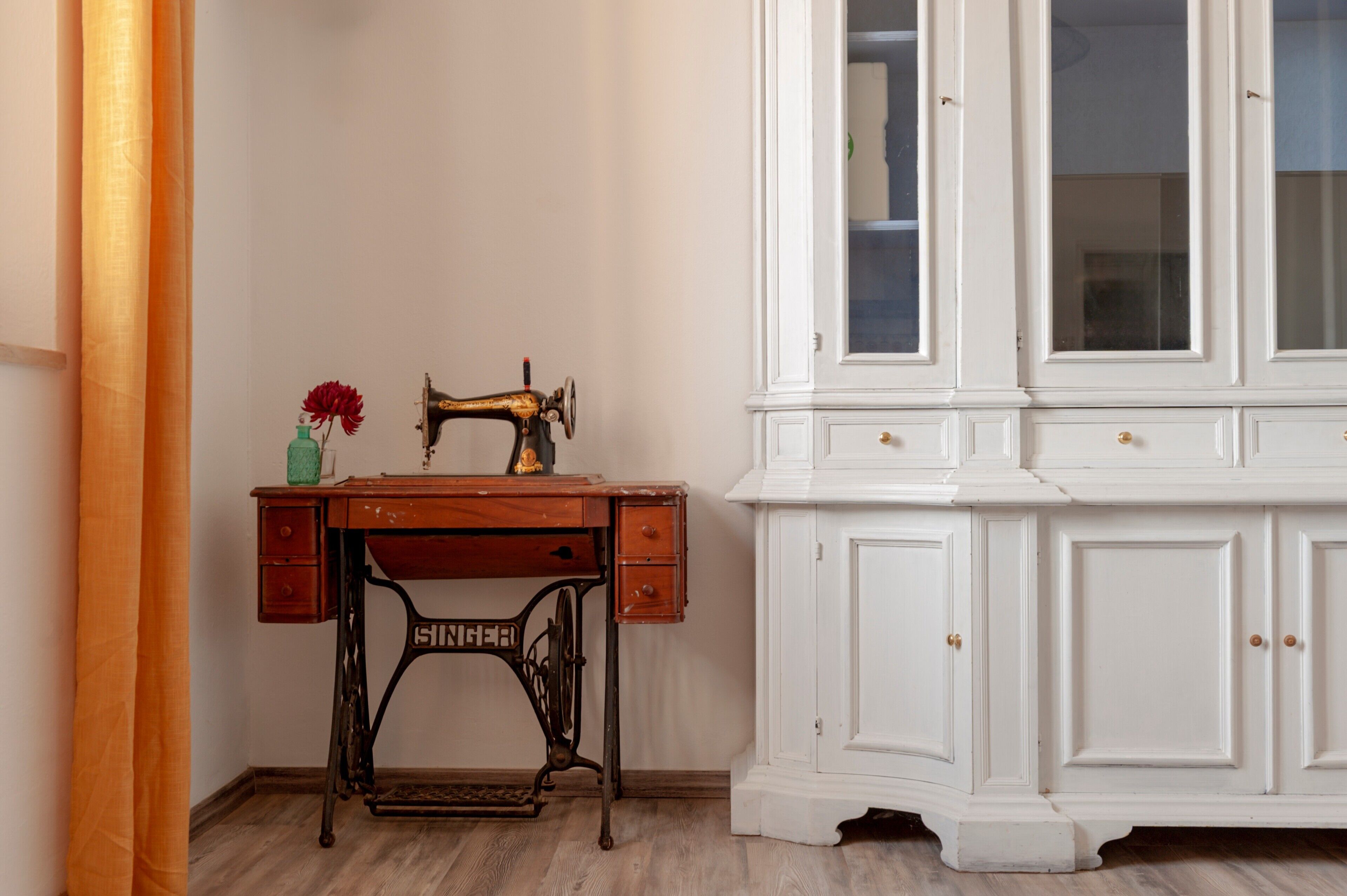 A room with a vintage sewing machine, a wooden desk, and a white cabinet with glass doors.