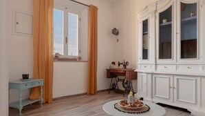 Une pièce avec un plancher en bois, une armoire blanche aux portes en verre, une machine à coudre, une petite table sur laquelle se trouve une assiette de nourriture et une fenêtre avec des rideaux.