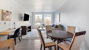 A modern living room with a dining area, a flat-screen TV mounted on the wall, and large windows providing natural light.