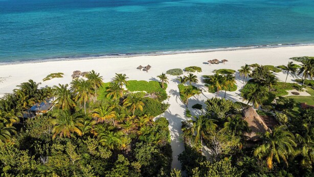 Luchtbeeld van een strandresort met palmbomen, een wit zandstrand en helderblauw water.