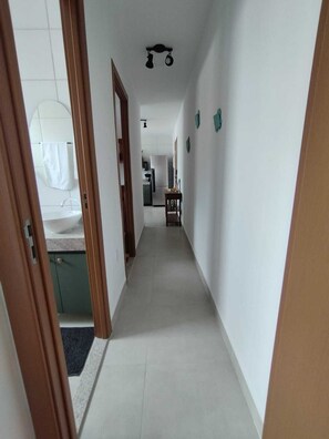 Um corredor estreito com chão de azulejos brancos, moldura de porta de madeira à esquerda e uma cozinha visível ao fundo.