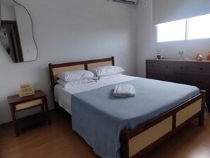 Un lit bien fait, recouvert d’une couvre-lit bleue et de draps blancs, une table de chevet en bois avec une lampe, une commode avec un miroir et une fenêtre laissant entrer la lumière naturelle.