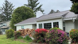 A single-story house with a front porch, a stack of firewood, and flowering bushes. - Charming Mill Beach Cottage by the Sea (Brookings)
