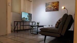 Ein Wohnzimmer mit Fernseher, einer Couch, einem kleinen Tisch mit Pflanze und einem Bild an der Wand.