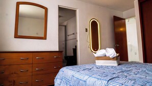 Ein Schlafzimmer mit einem hölzernen Schminktisch, einem Bett mit gestreifter Bettdecke, einem Spiegel mit Beleuchtung und einer Holztür.