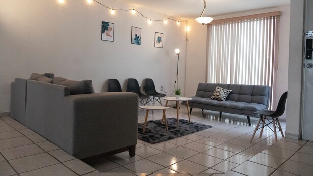 Ein modernes Wohnzimmer mit einem grauen Sofa, zwei schwarzen Sesseln, einem kleinen runden Tisch und einer Stehlampe. An der Wand hängen drei gerahmte Pflanzenmotive und eine Lichterkette ist darüber angebracht.