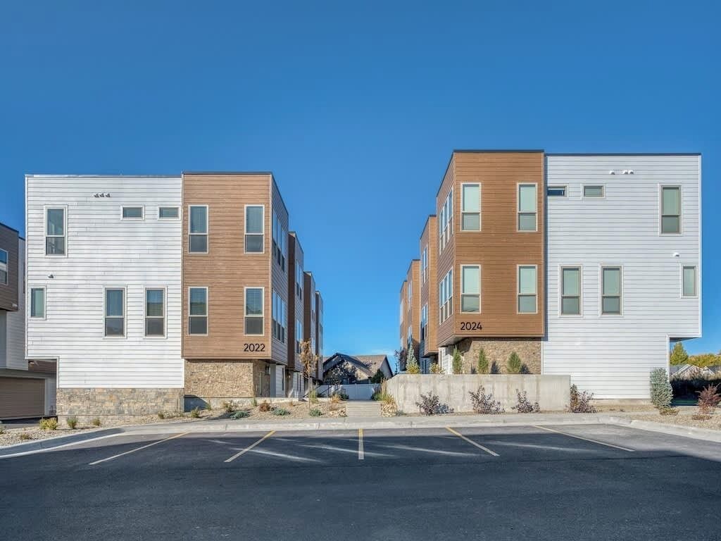 A modern apartment complex with two-story buildings, numbered 2022 and 2024, featuring a mix of wood and metal siding, and a parking lot in front.