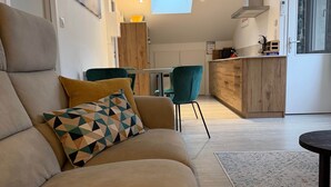 A modern living room with a sofa, a patterned throw pillow, a dining area with chairs, a kitchen with a window, and a skylight.