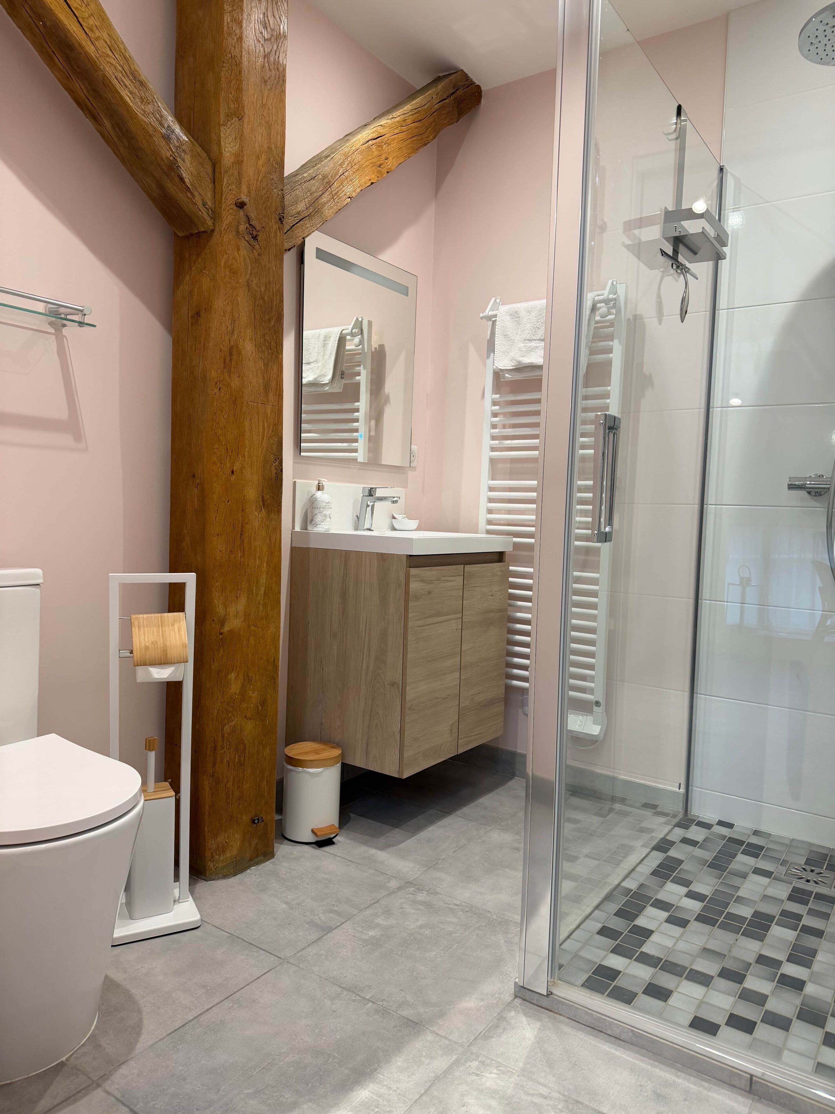 A bathroom with a toilet, sink, mirror, wooden beam, and a glass shower enclosure.