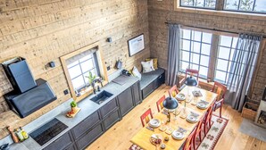 Una cocina moderna con una encimera alargada, un área de comedor con una mesa de madera y sillas rojas, y ventanas grandes con vista a árboles.
