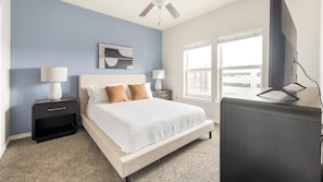A hotel room with a bed, two bedside tables with lamps, a ceiling fan, a TV, and a large window with a view. - Landing Apartments near Downtown Corpus Christi (Corpus Christi)