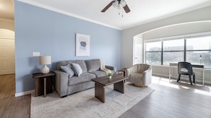 A living room with a grey sofa, a coffee table, a side table with a lamp, and a chair. There is a large window with a view of buildings. - Landing Apartments near Downtown Corpus Christi (Corpus Christi)