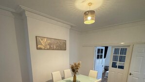 Een eethoek met een glazen tafel, witte stoelen, een vaas met gedroogde bloemen en een kroonluchter.