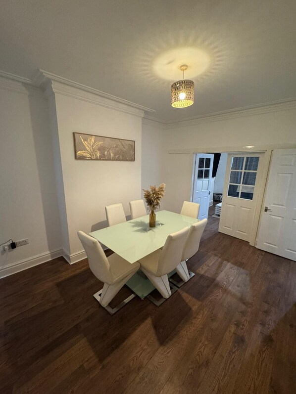 A dining area with a glass table, white chairs, a vase with dried flowers, and a chandelier.