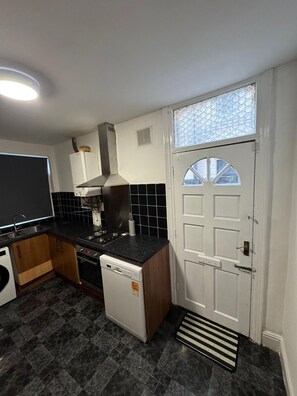 A compact kitchen with a stove, oven, dishwasher, and a small refrigerator. There is a window with a decorative grille and a white door with a circular window.