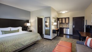 A hotel room with a large bed, a sitting area with a sofa and ottoman, a kitchenette, and a bathroom. - Studio Suite in Houston's Energy Corridor | Free Breakfast + 24h Business Center (Houston)