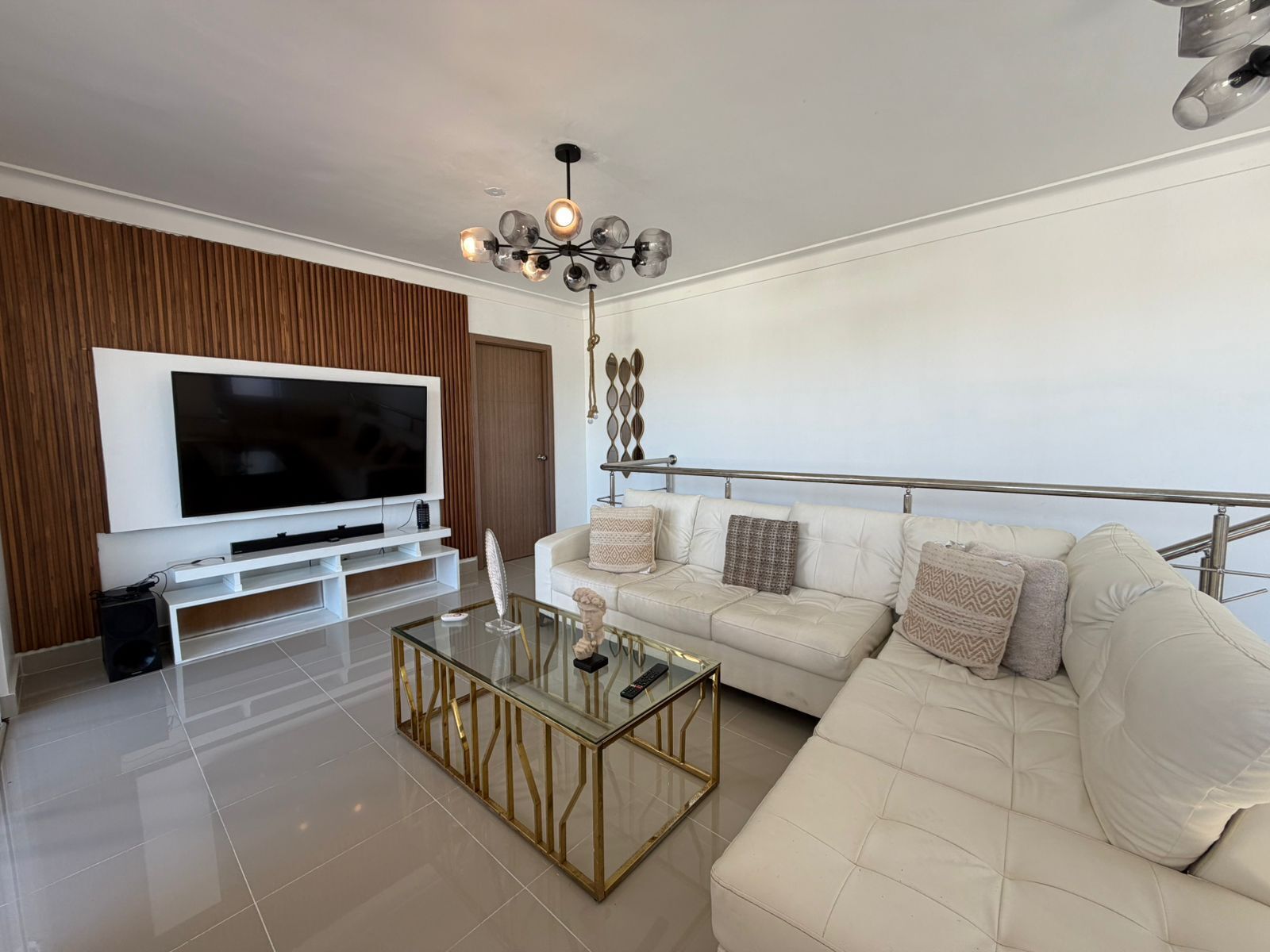 A modern living room with a large flat-screen TV, a glass coffee table, a white sofa, and a decorative ceiling light.