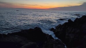 Paesaggio costiero con un tramonto sull'oceano, costa rocciosa e una piccola barca all'orizzonte.
