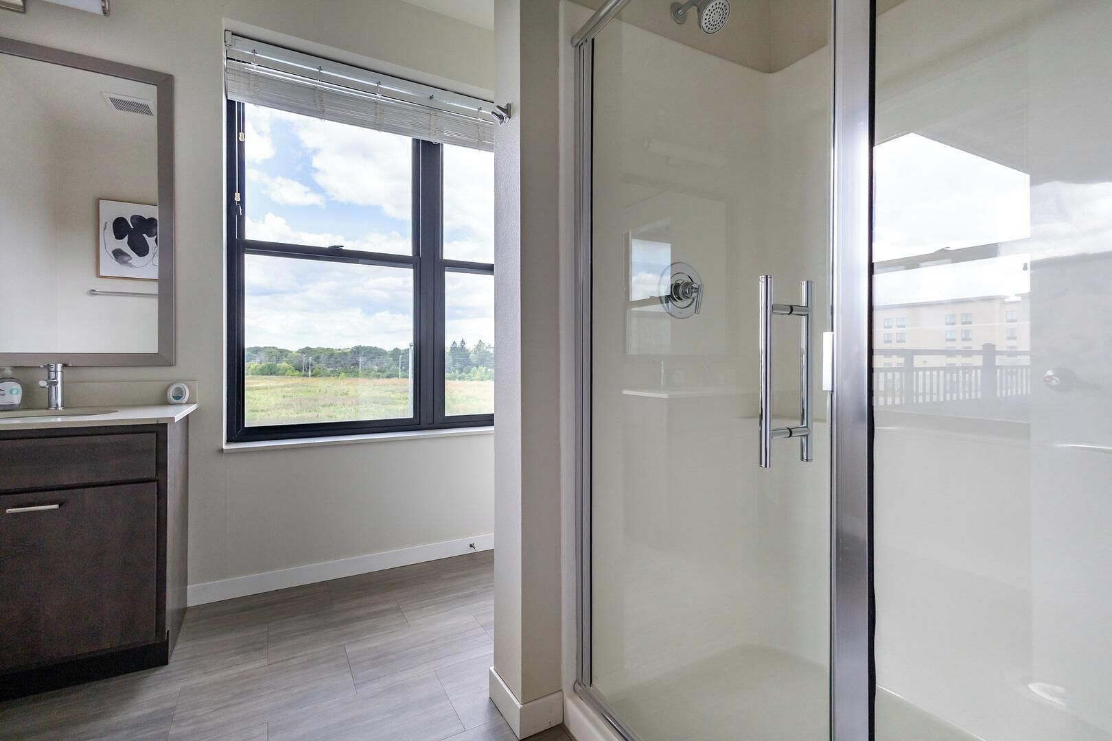 A modern bathroom with a glass shower enclosure, a large window with blinds, and a dark wooden vanity.
