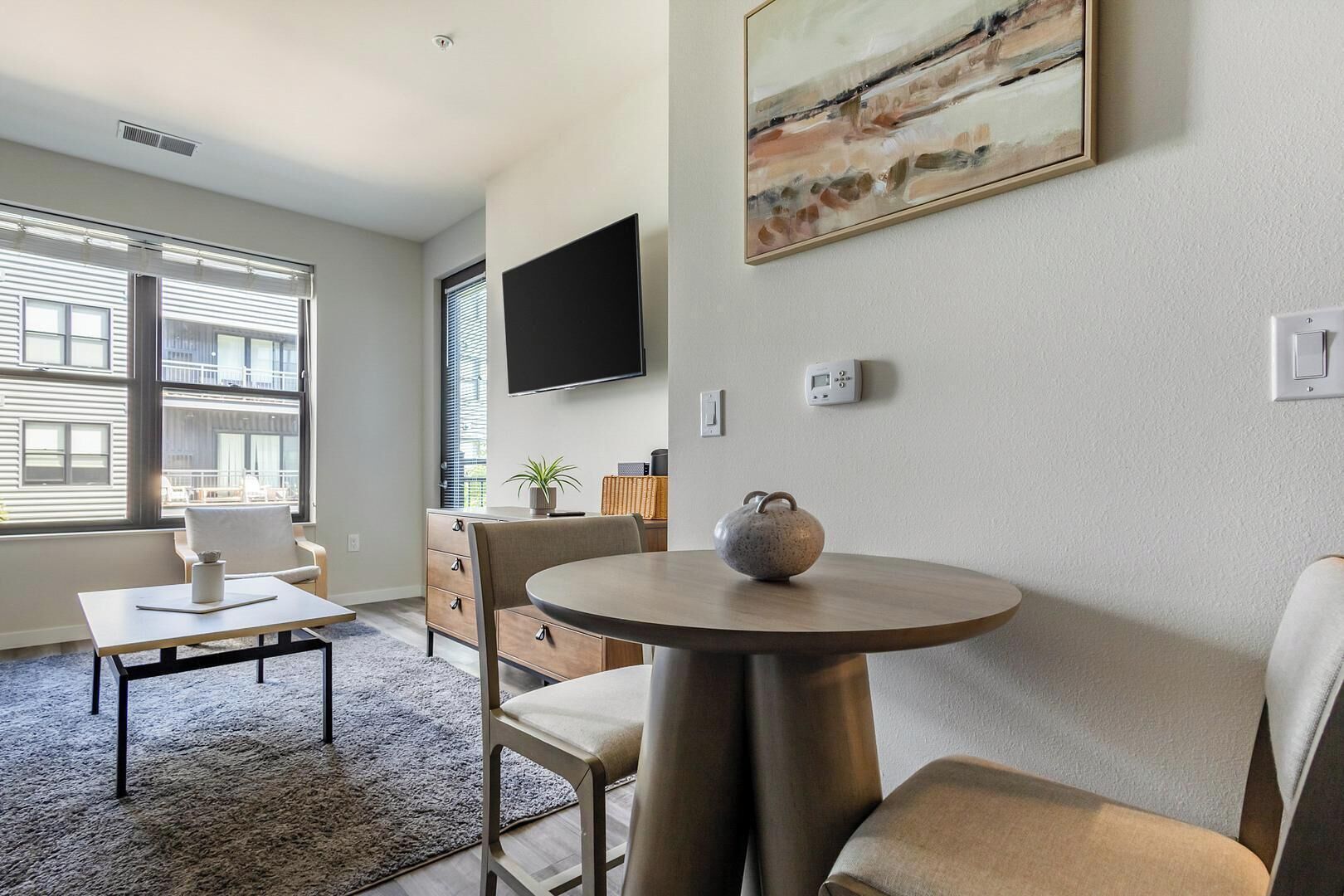 A modern living room with a round table, a flat-screen TV mounted on the wall, and a large window with blinds.