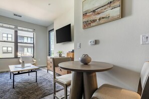 Een moderne woonkamer met een ronde tafel, een flatscreen televisie aan de muur en een groot raam met zonwering.