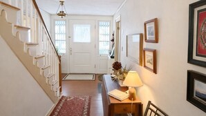 A hallway with a wooden staircase, a red carpet, a desk with a lamp, a chair with a book, and framed pictures on the wall.