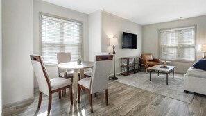 Ein modernes Wohnzimmer mit Essbereich, mit einem runden Tisch, zwei Stühlen, einer Couch, einem Couchtisch und einem an der Wand befestigten Fernseher.