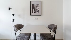 En stilren matplats med ett vitt bord och två stoppade stolar. En inramad, abstrakt tavla hänger ovanför bordet, och en modern golvlampa står placerad till vänster.