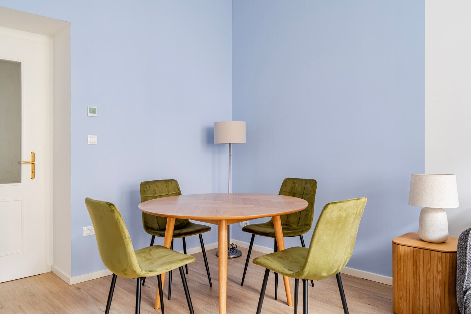 Une pièce avec une table ronde en bois entourée de quatre chaises vertes, une lampe sur une petite table d’appoint en bois et une porte avec une poignée dorée.