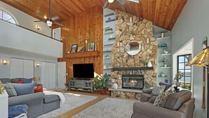 A spacious living room with a stone fireplace, wooden ceiling, and a large TV.