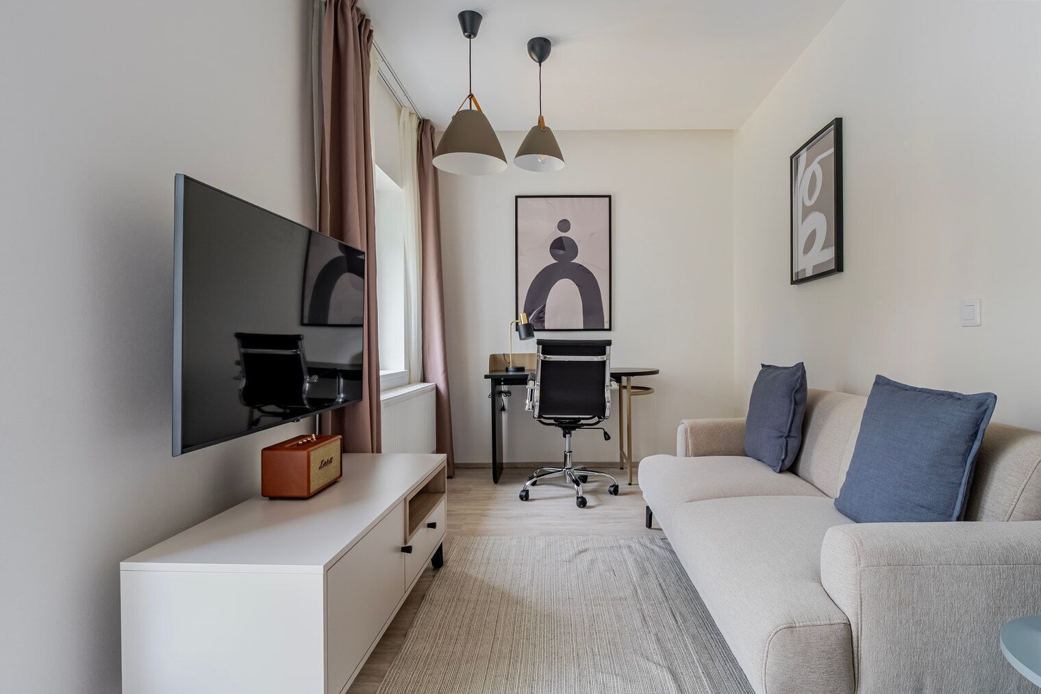 A modern hotel room with a flat-screen TV, a desk with a chair, a sofa with cushions, and a large window with curtains.