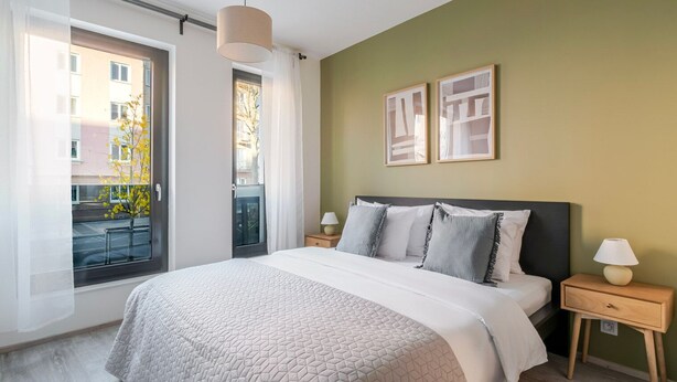 Ein Schlafzimmer mit einem großen Bett, einem Fenster mit Vorhängen, einem Nachttisch mit Lampe und zwei gerahmten Bildern an der Wand.