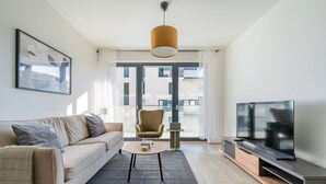 Ein modernes Wohnzimmer mit Sofa, Sessel, Couchtisch, Fernseher und einem großen Fenster mit Vorhängen.