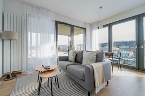 A modern living room with a grey sofa, a small round table, and a view of the city through large windows. - Blueground | Rohanský ostrov, balcony, Vltava view (PRG-91) (Prague)
